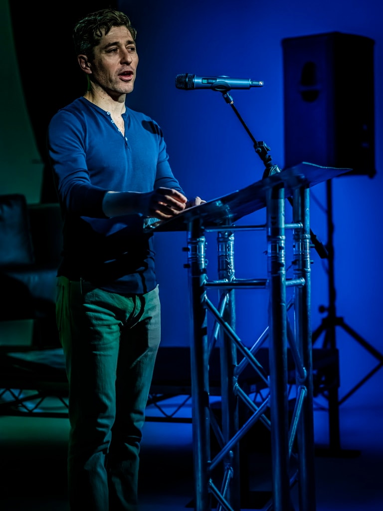 Jacob Frey speaking with papers in hand at the lectern, lit in blue stage light