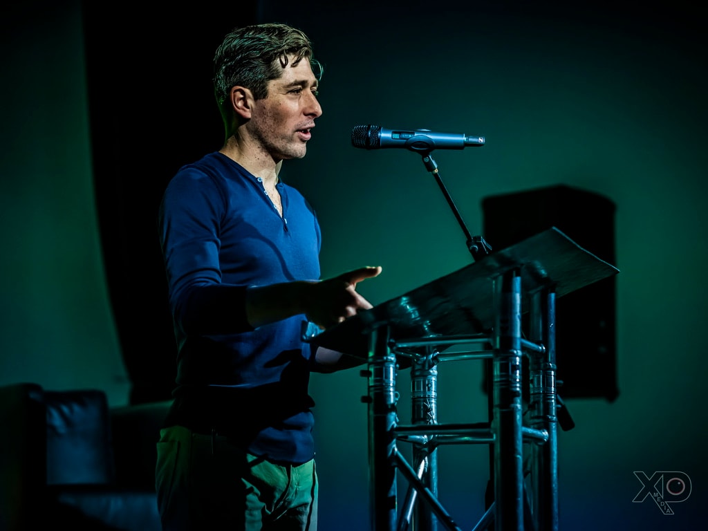 Jacob Frey in profile under green stage lighting, gesturing while speaking at a lectern
