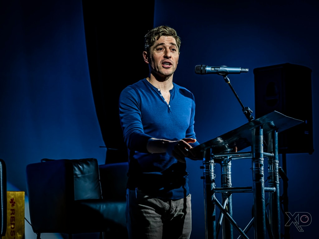 Jacob Frey speaking directly to the audience, blue stage lighting, venue signage visible in frame